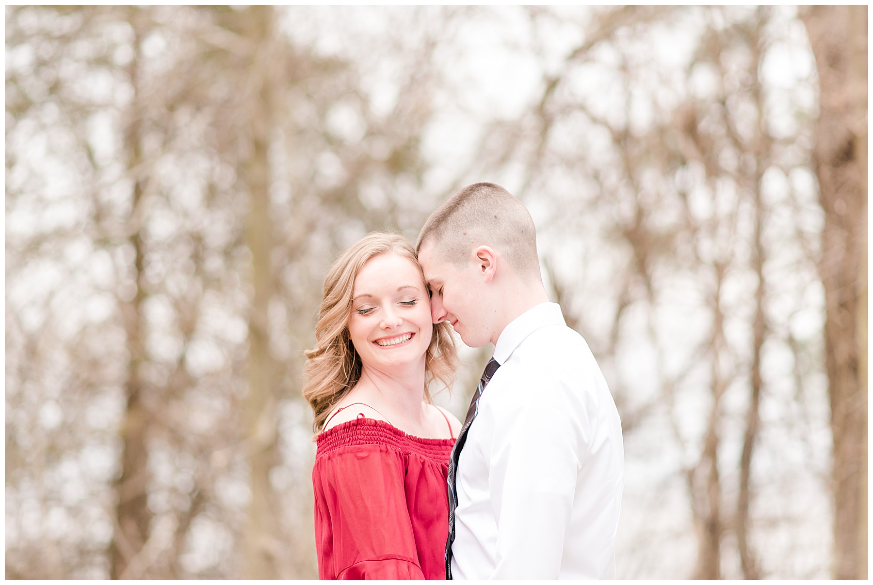 Bride in a red dress is chest to chest with the groom. He is nuzzling her temple while she in smiling with her eyes softly closed for an engagement session at Glenns Valley Nature Park