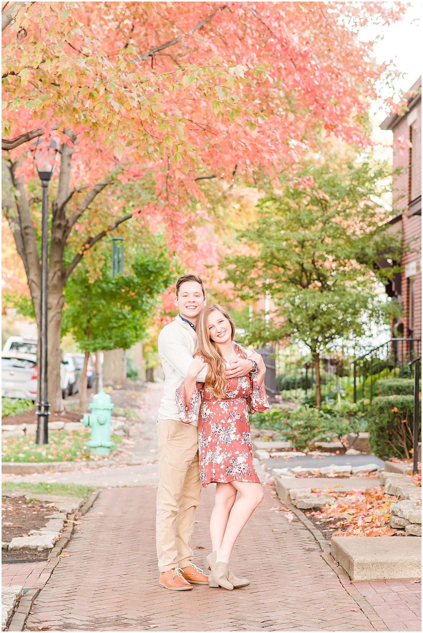 Couple hugging and smiling at camera in front of red trees during Lockerbie Square engagement session