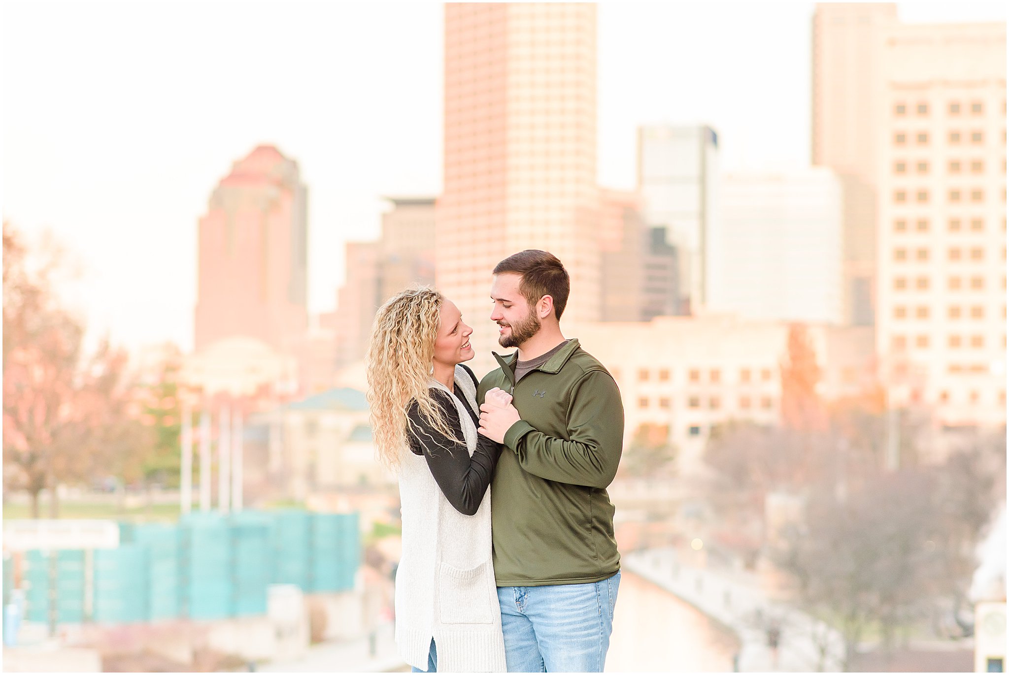 Couple holding hands and smiling at each other in front of Indianapolis skyline during Indianapolis Canal Engagement Session