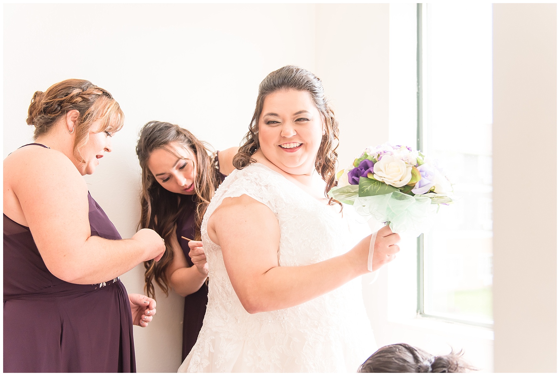 Bride smiling over shoulder as bridesmaids button her lace wedding dress